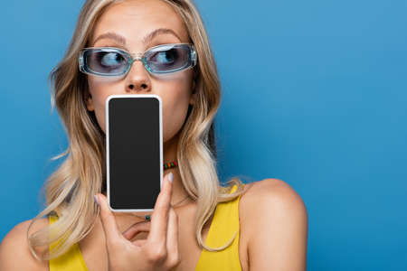 young blonde woman in sunglasses covering mouth while holding smartphone with blank screen isolated on blueの写真素材