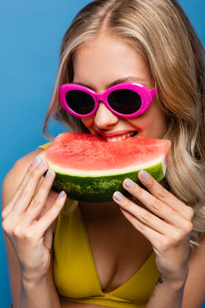 cheerful young woman in pink sunglasses eating fresh watermelon isolated on blueの写真素材