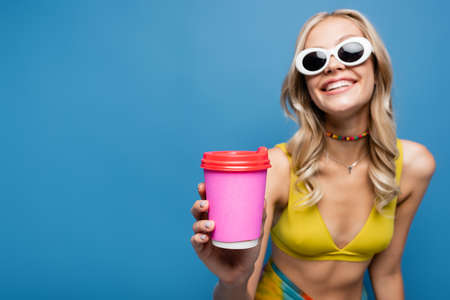 happy young woman in sunglasses and bikini top holding paper cup isolated on blueの写真素材
