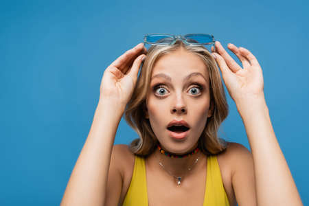 shocked young woman adjusting trendy sunglasses and looking at camera isolated on blueの写真素材