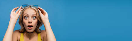 shocked young woman adjusting trendy sunglasses and looking at camera isolated on blue, banner