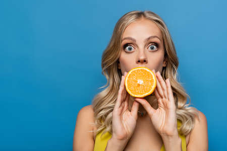 shocked young woman covering mouth with orange half isolated on blueの写真素材