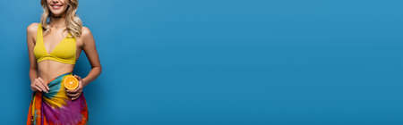 cropped view of cheerful young woman in yellow bikini top and sarong holding orange half on blue, bannerの写真素材