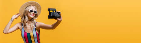 joyful woman in straw hat and swimsuit talking selfie on vintage camera isolated on yellow, bannerの写真素材