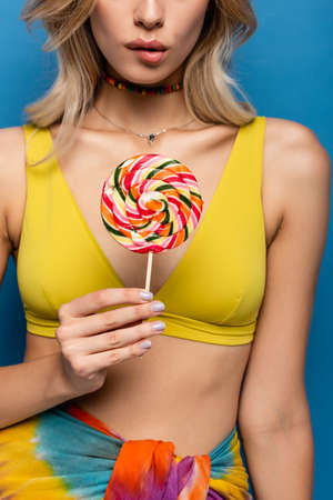 cropped view of young woman in yellow bikini top holding sweet lollipop isolated on blue