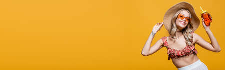 smiling woman in straw hat and sunglasses holding alcohol cocktail isolated on yellow, banner