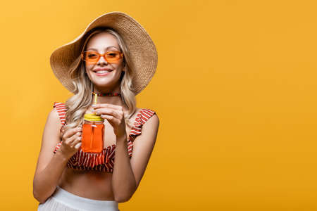 cheerful woman in straw hat and sunglasses holding alcohol cocktail isolated on yellow