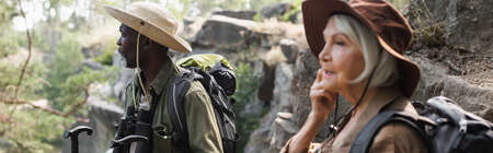 Senior african american tourist with trekking poles standing near blurred wife in forest, bannerの写真素材