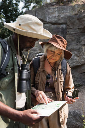 Senior hiker with compass pointing at map near african american husband with binocularsの写真素材