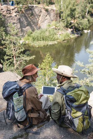 Senior woman with backpack looking at african american husband with digital tablet near lakeの写真素材