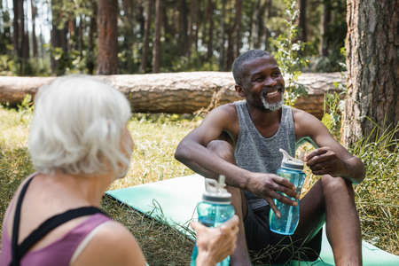 African american sportsman holding sports bottle near blurred wife in forest