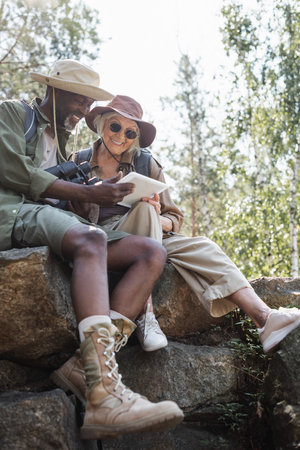 Smiling multiethnic hikers with binoculars and digital tablet sitting on stones in forestの写真素材