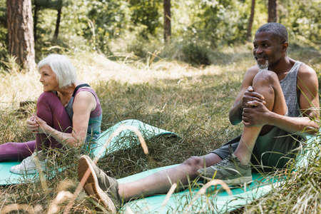 Smiling african american man stretching on fitness mat near wife in forestの写真素材