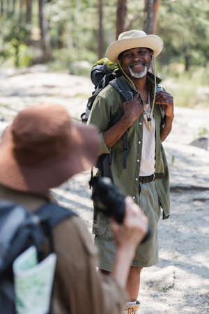 Cheerful african american tourist with backpack looking at blurred wife in forest