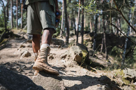 Cropped view of african american traveler holding binoculars on path with stones in forest