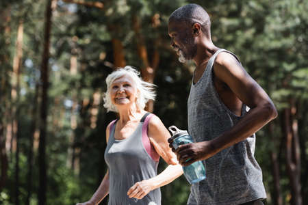 African american sportsman holding sports bottle while jogging with wife in forestの写真素材