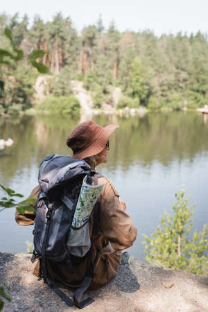 Elderly hiker with map and backpack sitting on cliff near lakeの写真素材