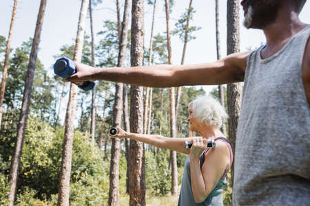 Side view of elderly woman training with dumbbells near blurred african american husband in forest