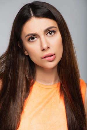 pensive young woman biting lip and looking at camera isolated on gray