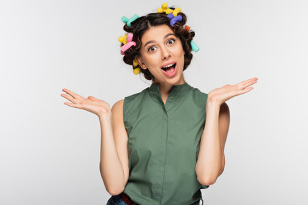 excited young woman with colorful hair curlers gesturing isolated on gray