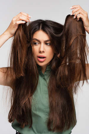 dissatisfied young woman looking at tangled long hair isolated on gray