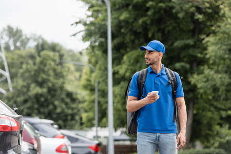 Smiling muslim deliveryman with backpack and smartphone walking near cars outdoorsの写真素材