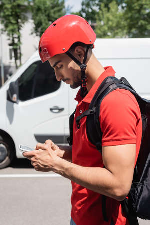 Side view of arabian courier in helmet using smartphone on urban streetの写真素材
