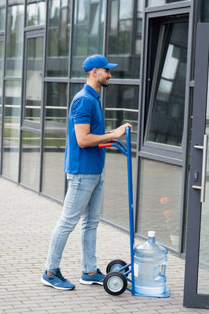 Side view of smiling muslim courier standing near bottle of water on cart and buildingの写真素材