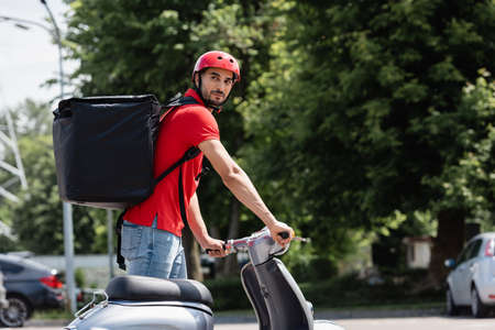 Young arabian deliveryman with thermo backpack standing near scooter on urban streetの写真素材