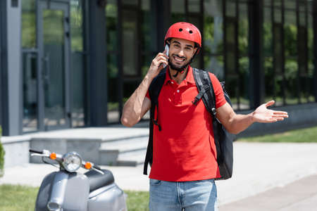 Cheerful muslim courier with thermo backpack talking on smartphone near blurred scooter outdoorsの写真素材