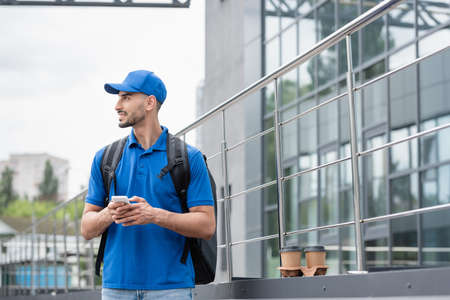 Side view of smiling arabian courier with thermo backpack holding mobile phone near paper cup and building outdoorsの写真素材