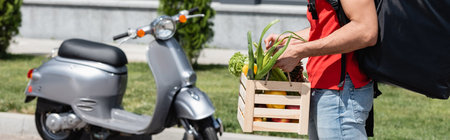 Cropped view of deliveryman with vegetables and backpack standing near blurred scooter outdoors, bannerの写真素材