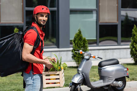 Arabian courier with vegetables and thermo backpack smiling at camera near blurred scooterの写真素材