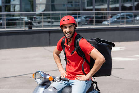 Excited arabian deliveryman with thermo backpack holding hands on hips while sitting on scooterの写真素材