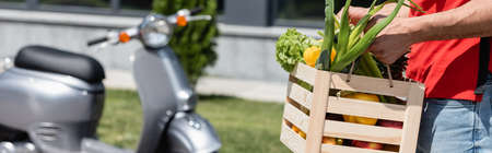 Cropped view of courier holding box with ripe vegetables near blurred scooter, bannerの写真素材