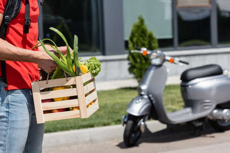 Cropped view of deliveryman holding wooden box with fresh vegetables near blurred scooter outdoorsの写真素材