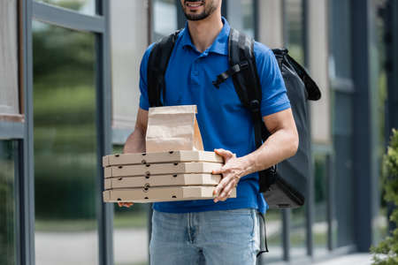 Cropped view of cheerful deliveryman holding pizza boxes and paper bag near building