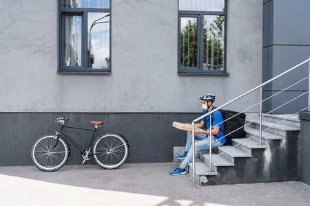 Arabian deliveryman in medical mask holding pizza boxes near bike and building outdoorsの写真素材