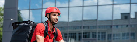 Cheerful muslim courier in helmet and thermo backpack outdoors, bannerの写真素材