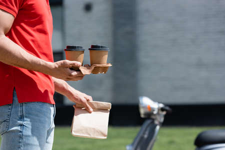 Cropped view of courier holding coffee to go and paper bag near blurred scooter outdoorsの写真素材