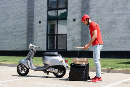 Side view of young muslim deliveryman in helmet holding pizza box near thermo backpack and scooterの写真素材