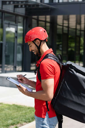 Side view of cheerful arabian courier with thermo backpack and clipboard standing outdoorsの写真素材