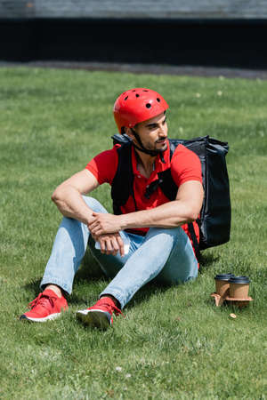 Young muslim courier in helmet and thermo backpack holding smartphone near paper cups on grassの写真素材