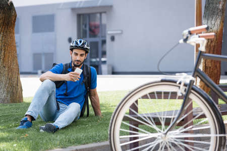 Arabian courier in helmet using smartphone on lawn near blurred bike on urban streetの写真素材