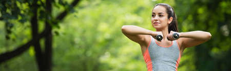 young sportswoman looking away while working out with dumbbells in park, bannerの写真素材