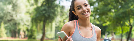 happy sportswoman holding sports bottle and smartphone in park, bannerの写真素材