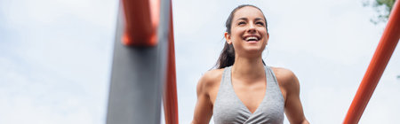 low angle view of cheerful and fit woman in sportswear exercising on parallel bars outside, banner