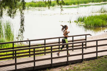 full length of happy woman in crop top and leggings jogging on bridge near lake in parkの写真素材