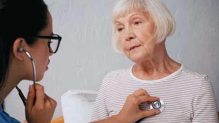 blurred nurse in eyeglasses examining aged woman with stethoscopeの写真素材