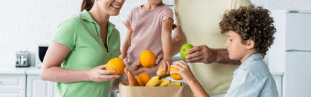 Smiling woman holding fruit near family and paper bag in kitchen, bannerの写真素材